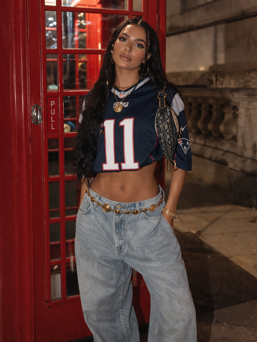 Woman in bold urban sporty chic outfit 2026 wearing a cropped navy NFL jersey with number 11, light-wash baggy wide-leg jeans, gold coin belt, layered gold necklaces, large hoop earrings, and a small Dior saddle bag, standing in front of a red London telephone booth at night