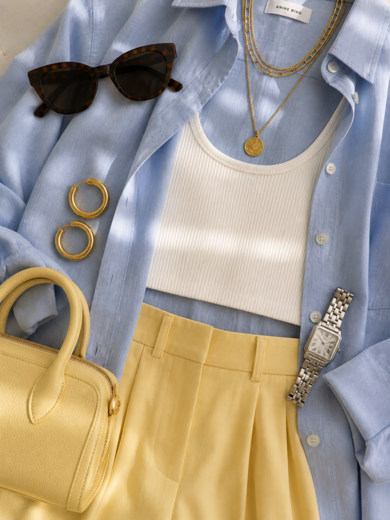 Close-up detail of clean chic effortless outfit 2026 showing chunky gold hoops, layered gold necklaces, mini butter yellow handbag, silver watch, cat-eye sunglasses, and soft girl perfume alongside the blue shirt and yellow trousers